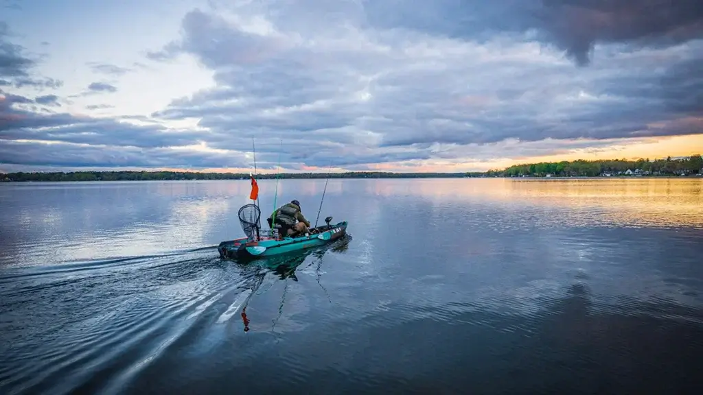 A view from behind a fishing kayak moving across a lake, equipped with a stern-mounted trolling motor, rod holders, and an angler, under a cloudy sky at dusk.