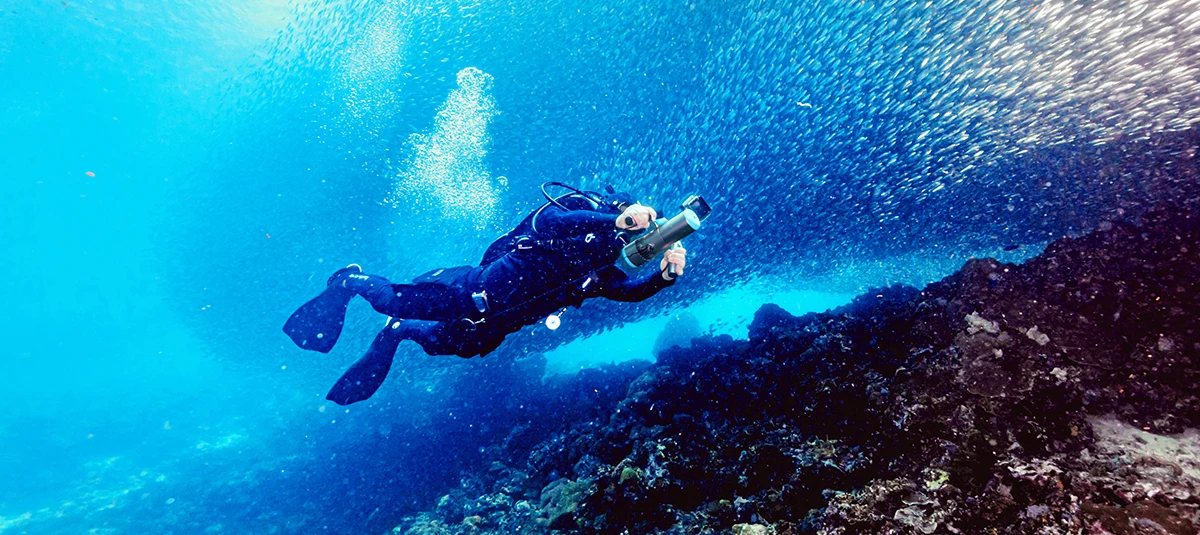 Scuba diver exploring a coral reef with TEDGIX K5 underwater scooter, swimming effortlessly alongside a massive school of fish in the deep ocean.